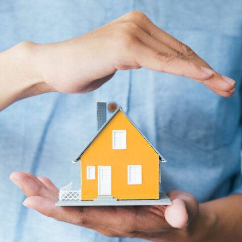 Person hält schützend Hand über ein Modellhaus
